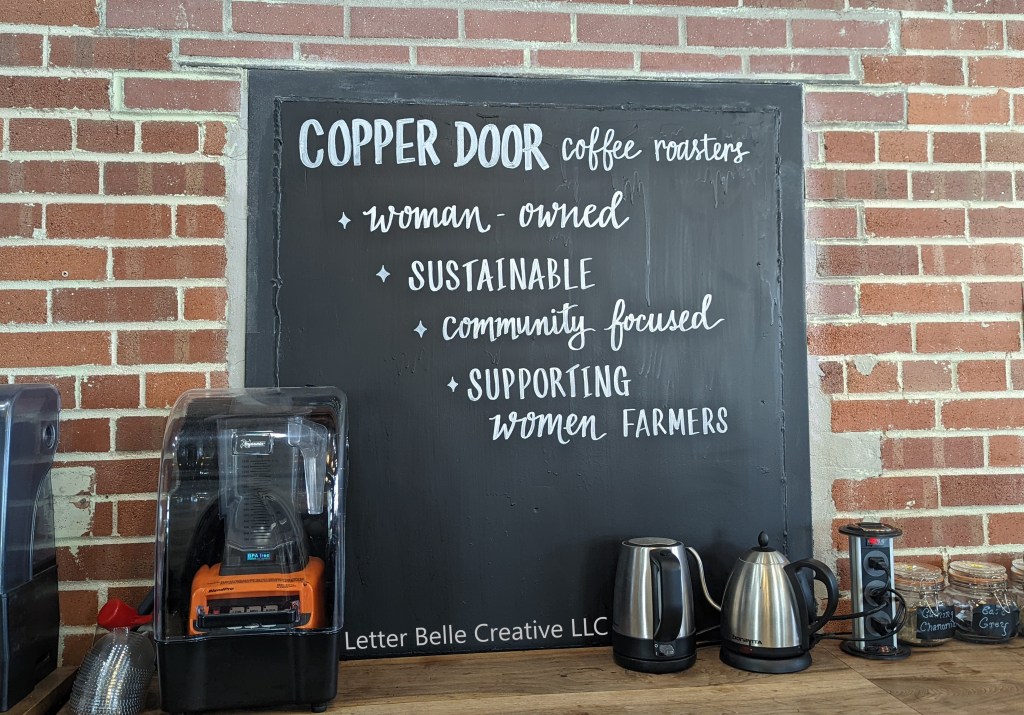 Black chalkboard embedded in brick wall at Copper Door Coffee Roasters in Lowry. White hand lettering: Copper Door Coffee Roasters -woman-owned -sustainable -community focused -supporting women farmers

Letter Belle Creative