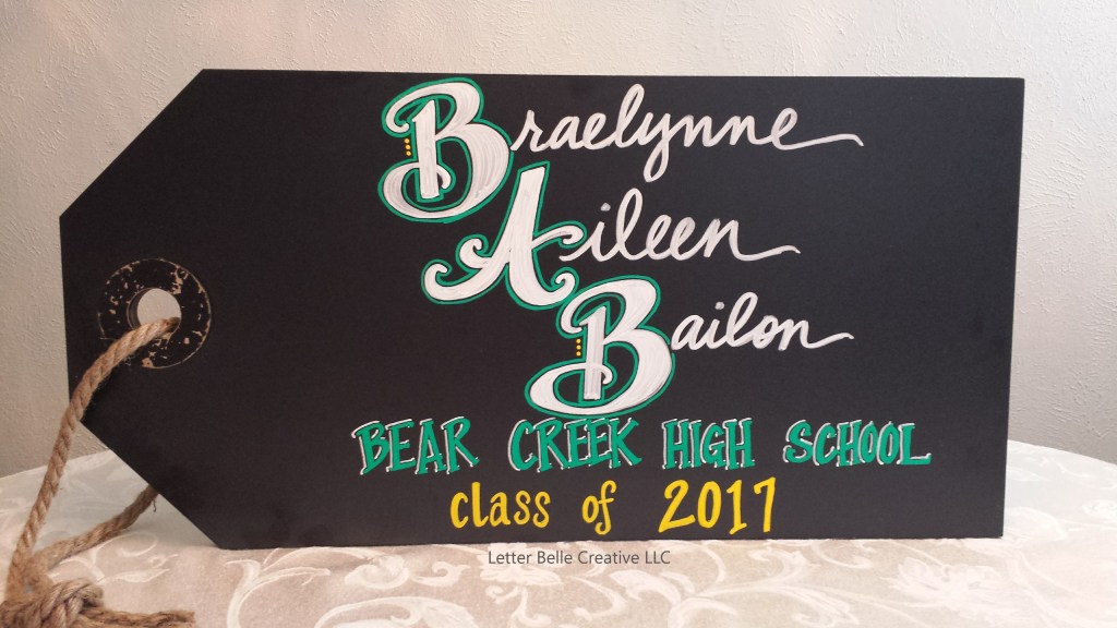 Black chalkboard in shape of large gift tag. White, green & yellow writing: full name, high school name, class of 2017. Used as prop in Senior photos.

Letter Belle Creative
