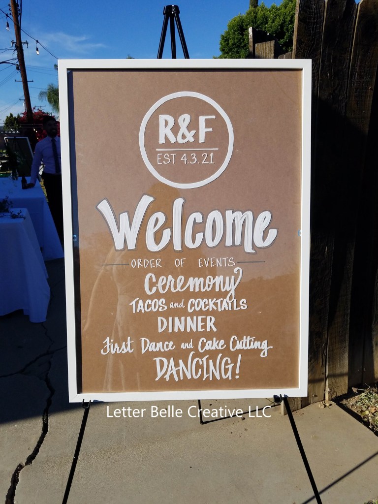 Framed acrylic used for welcome & order of events sign at wedding. Write writing: R&F est 4.3.21. Welcome. Order of events: ceremony, tacos & cocktails, dinner, first dance & cake cutting, dancing!

Letter Belle Creative